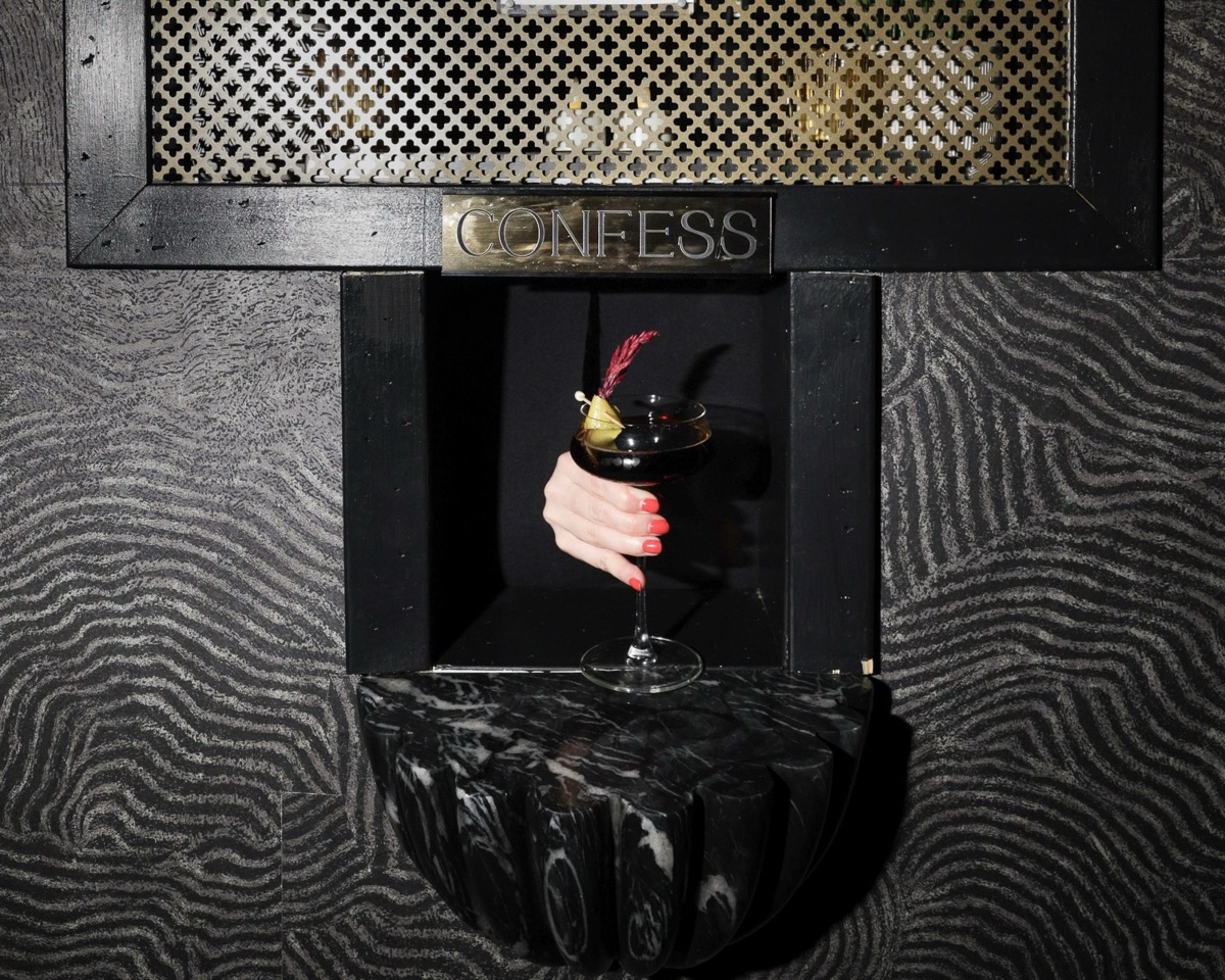 The CONFESS window — a brass grille and plaque over a black marble ledge holding a cocktail
